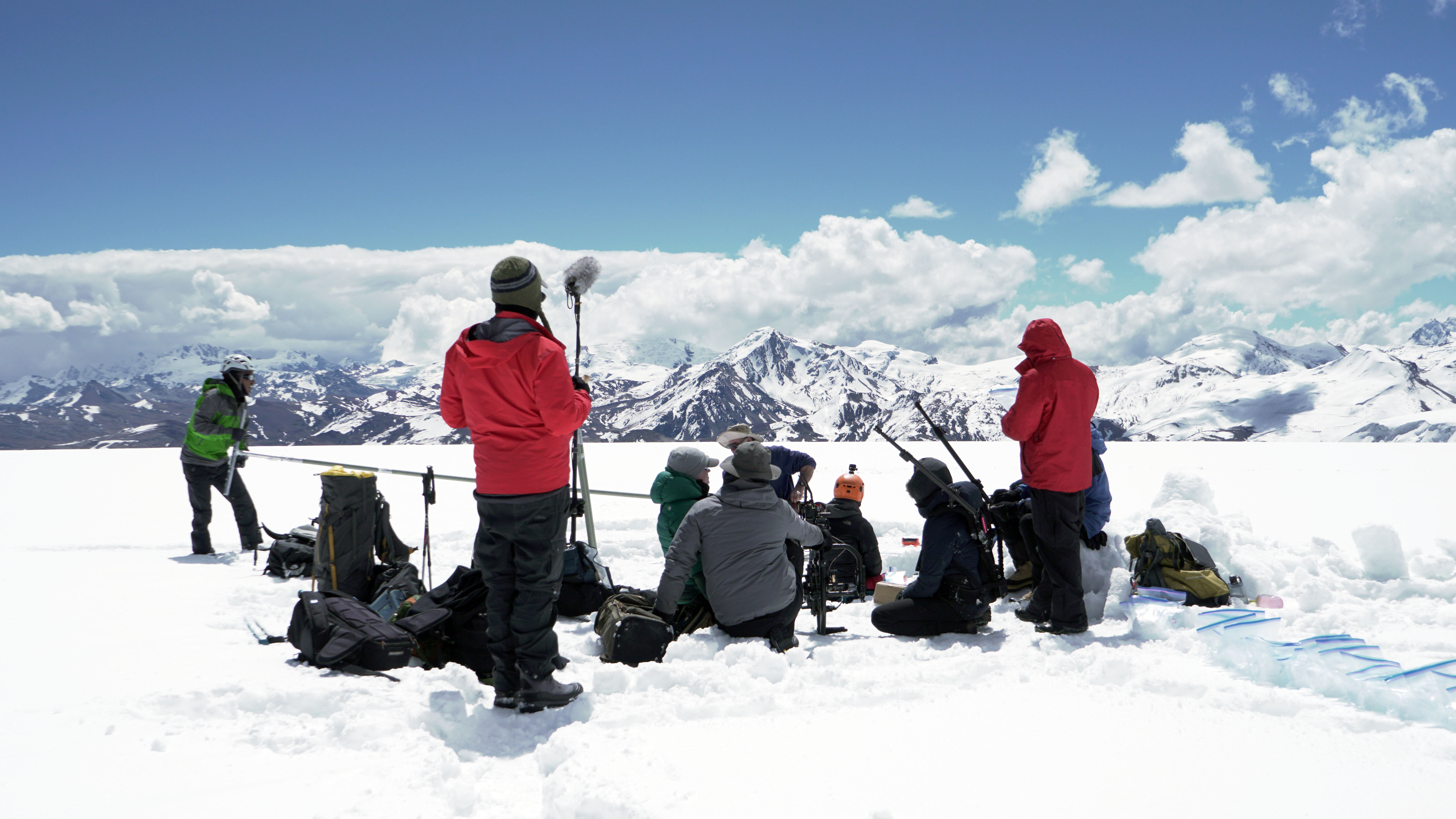 A photo of a crew of people sitting and standing in snow with filming equipment and with mountains in the background