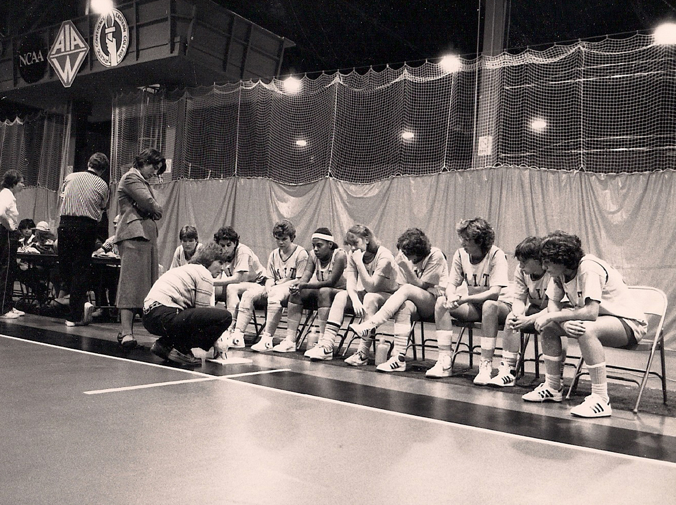 A black and white photo inside a basketball court with a basketball team sitting in chairs in a line with one coach standing and one crouching on the sidelines talking to the team