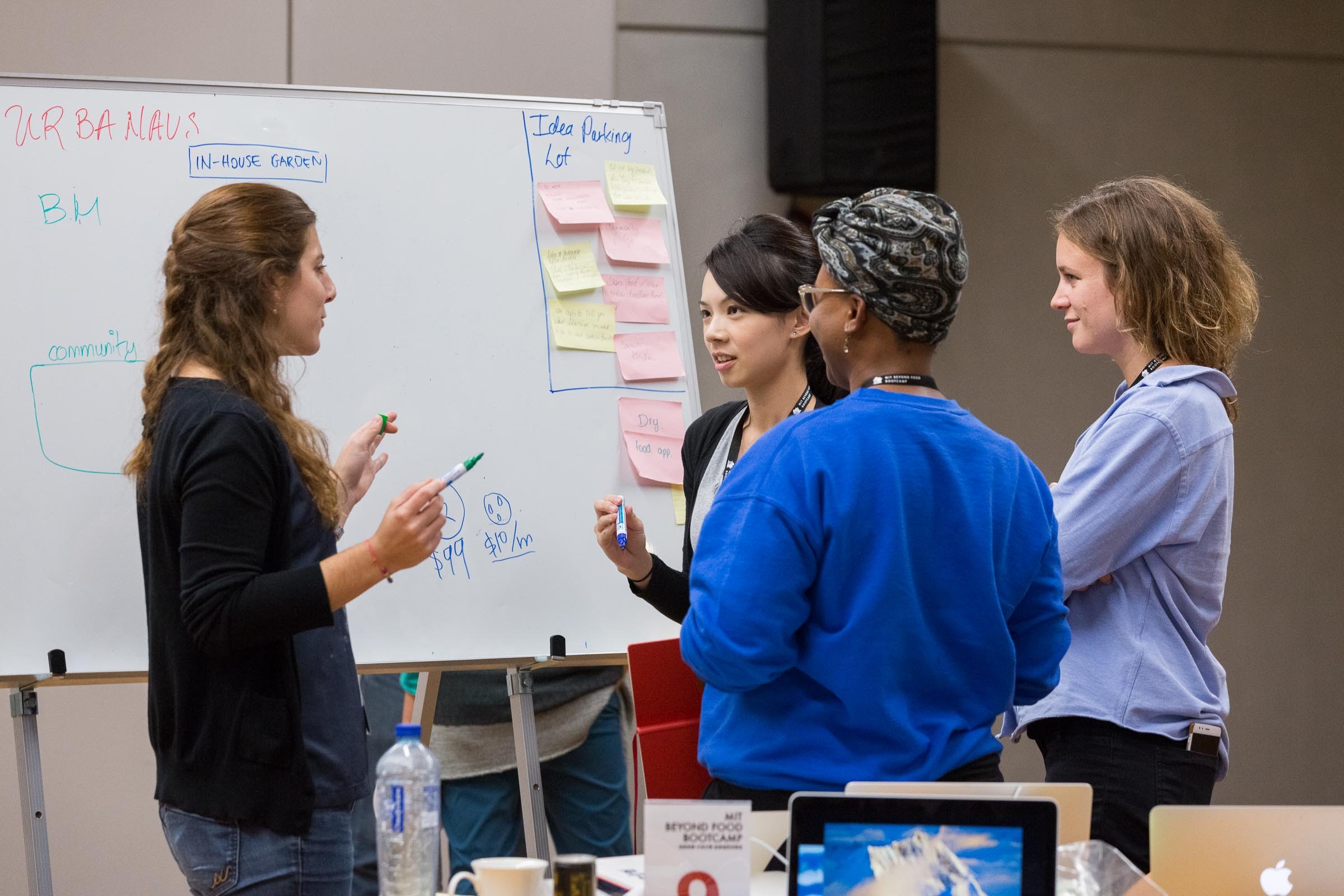 Four women converse in front of a white board covered with post-its.