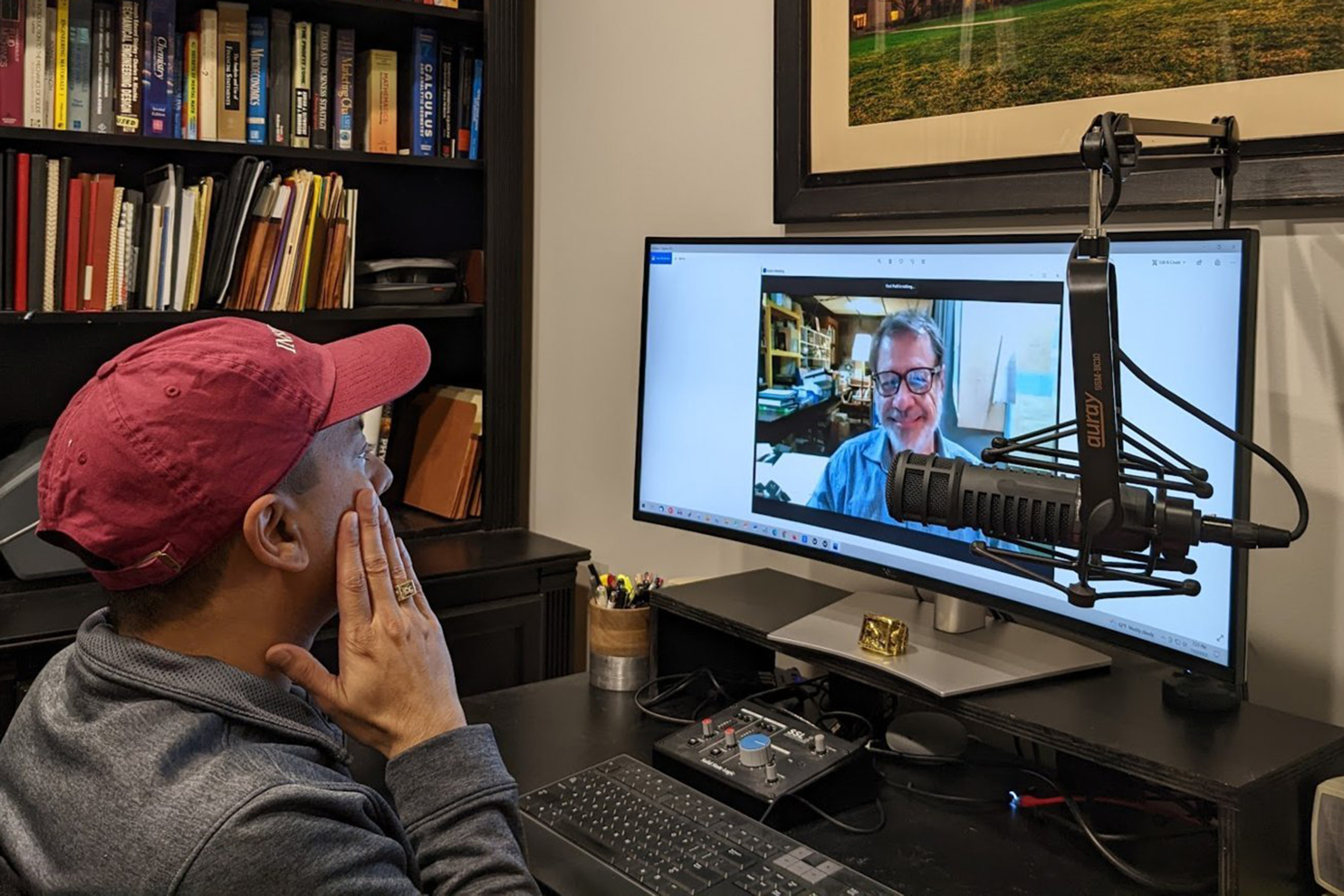 Profile view of Ravi Patil from shoulders up sitting in an office looking at a screen. He wears a red ballcap and has his hand to his cheek. A microphone partly obscures the screen, which shows a head and shoulders view of Curtis Blaine in an office.