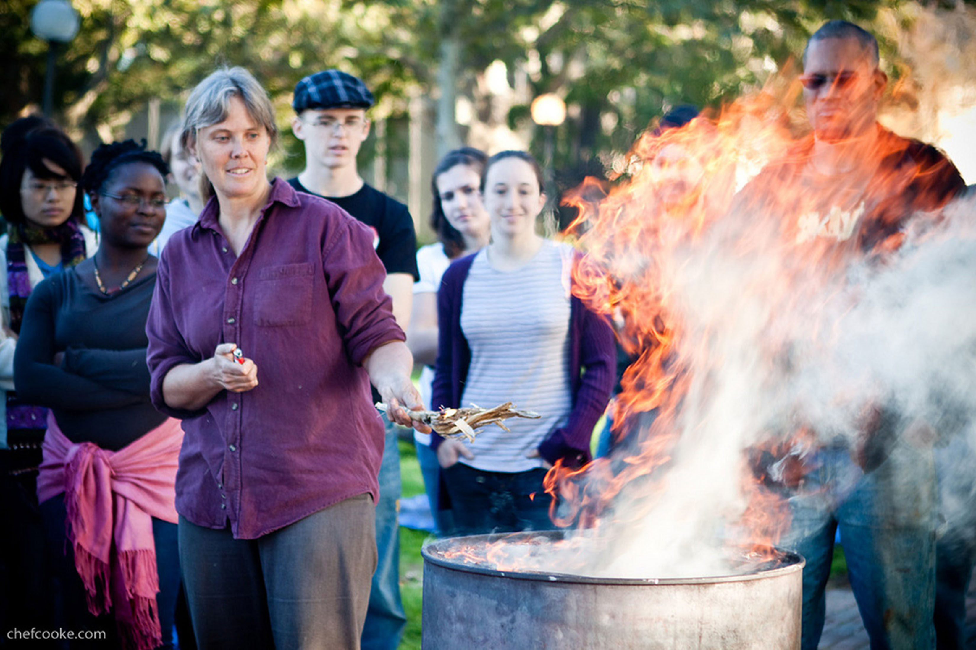 Amy Smith stands next to a trashcan holding a lighter and some sticks. Flames are pouring from the trashcan. Several people stand behind Smith, watching. 