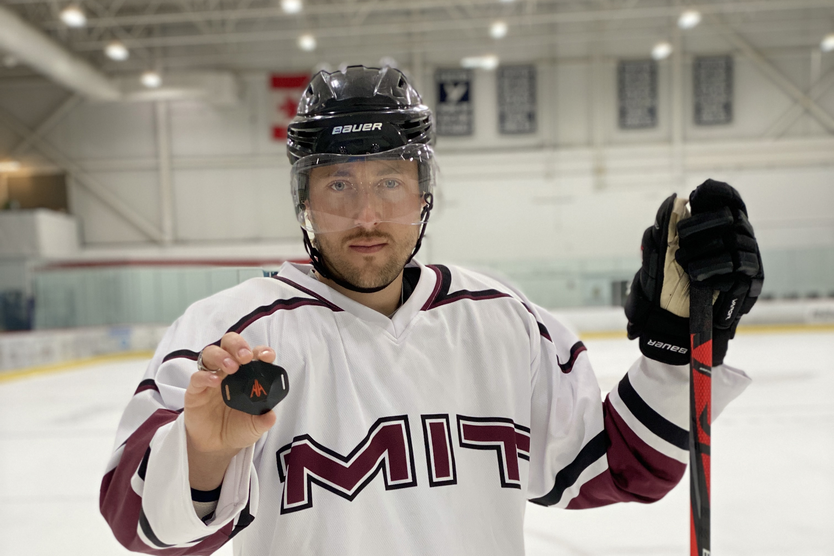 Photo of Bill Near standing in an ice rink wearing an MIT jersey and holding a black plastic device in his right hand and a hockey glove and stick in his left