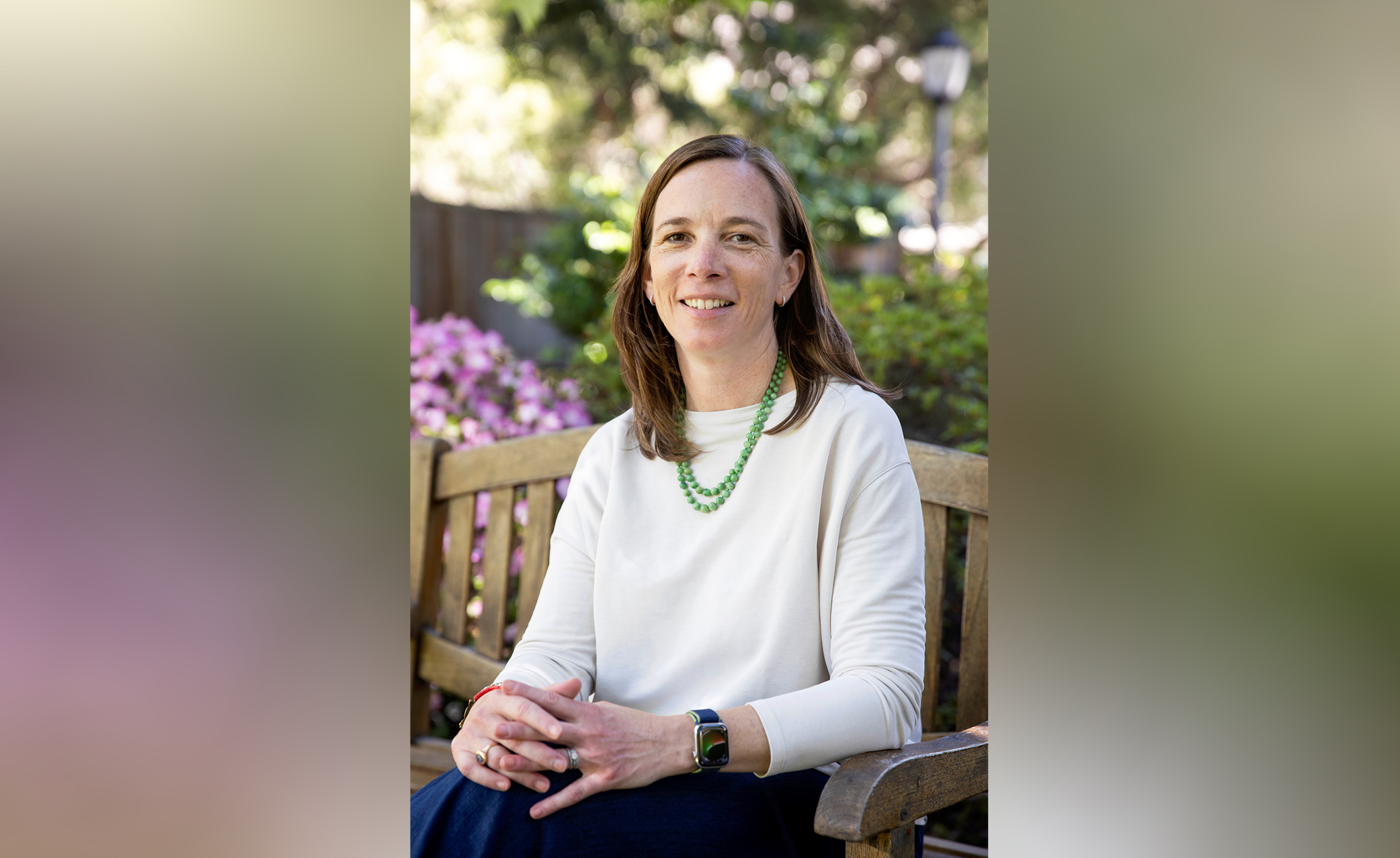 Louise Bedsworth sitting on a wooden bench outside with pink flowers to the left and green and trees behind and overhead