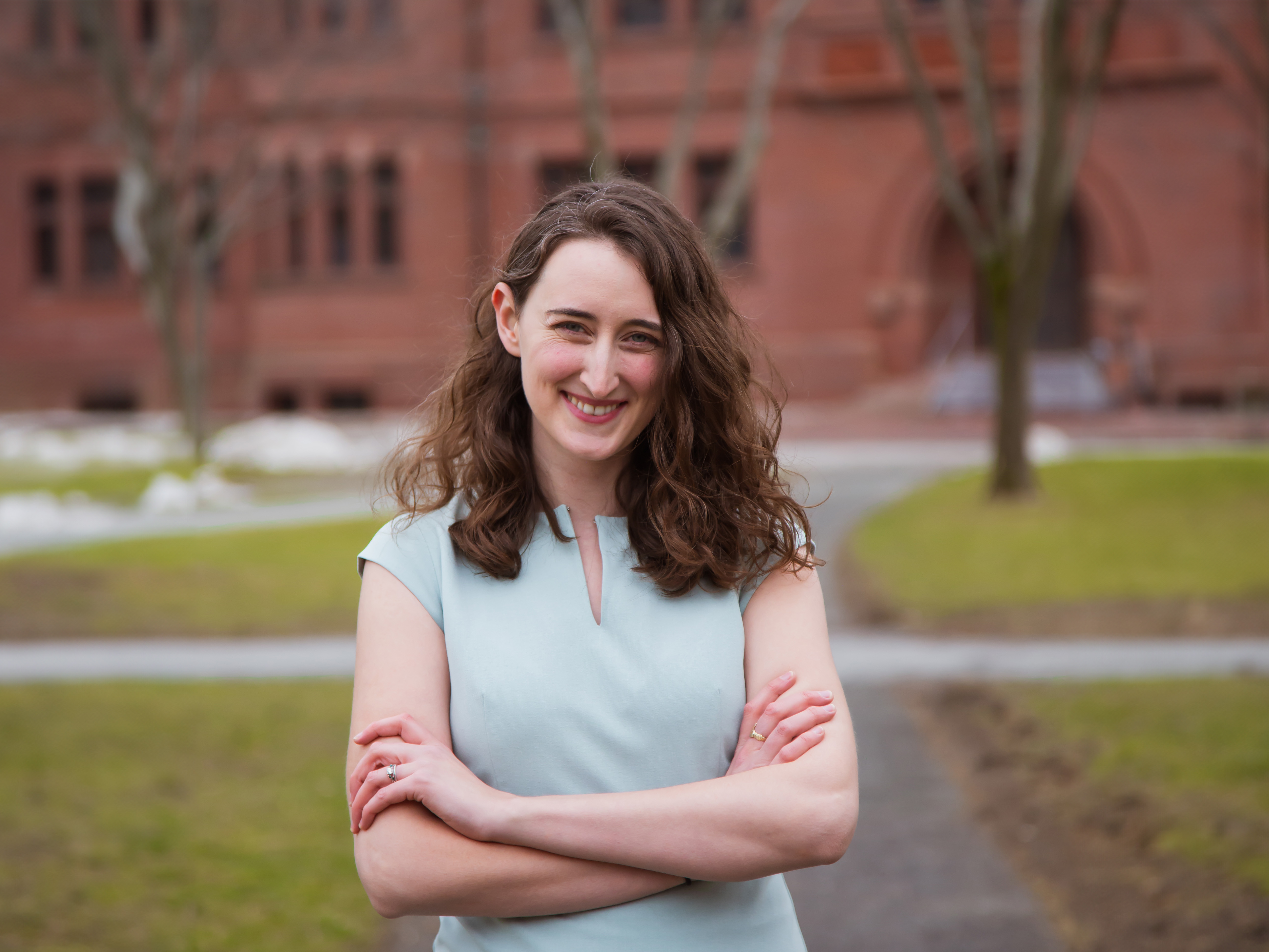 Photo of Cassandra Zentner standing on a walkway outside between grassy patches with her arms crossed and a brick building behind her