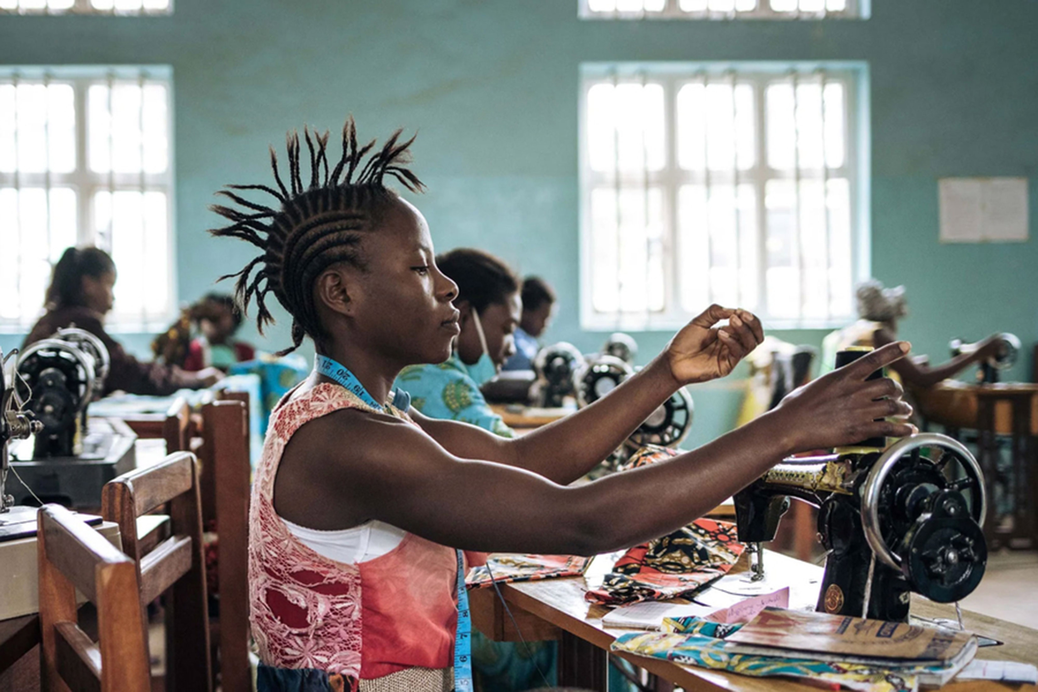 A woman threads a sewing machine in a room with other sewers. There are colorful fabrics on her sewing table.