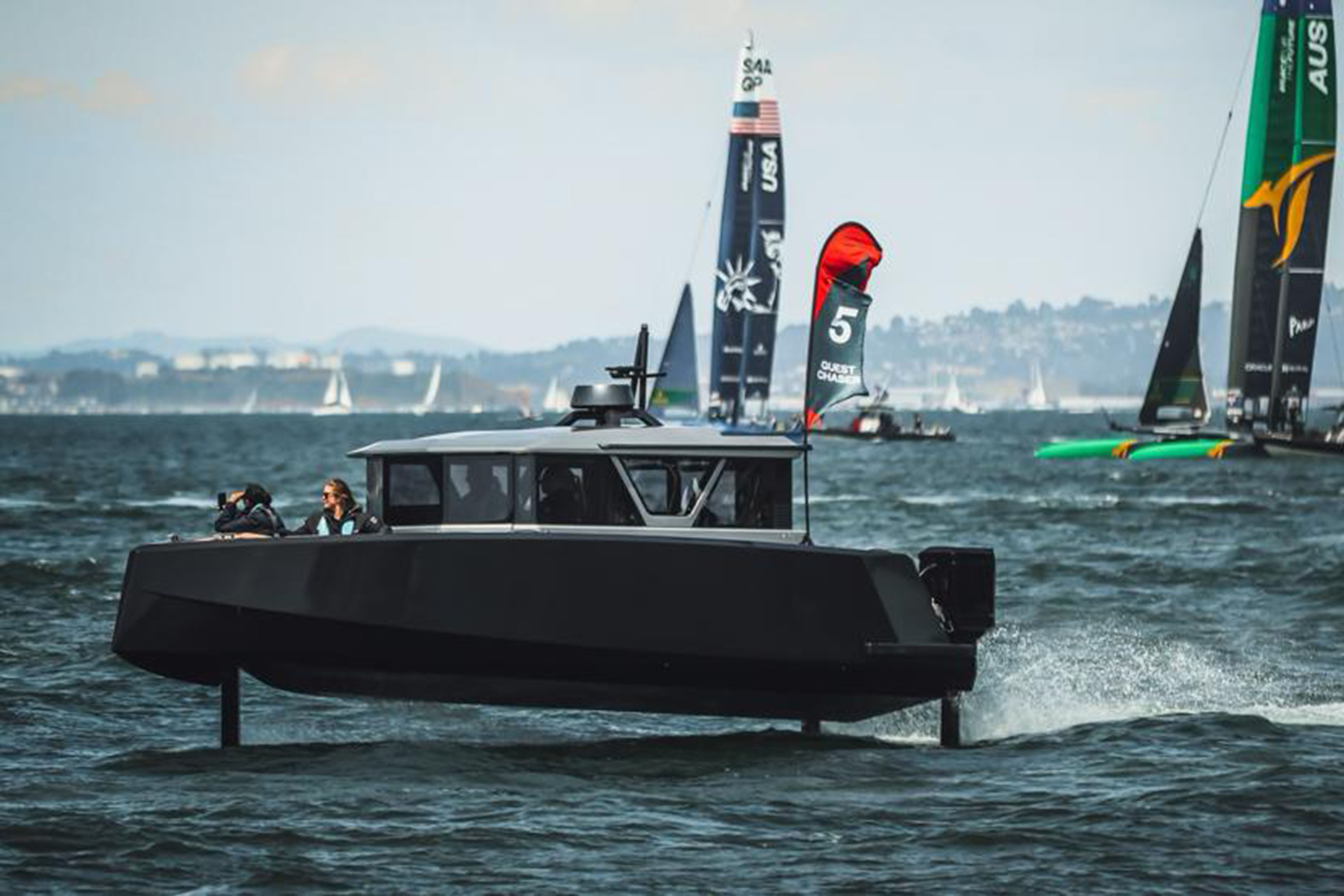 A black boat appears on the open water with sailboats in the background. The body of the boat is above the water's surface. Three stilts hang down from it into the water.