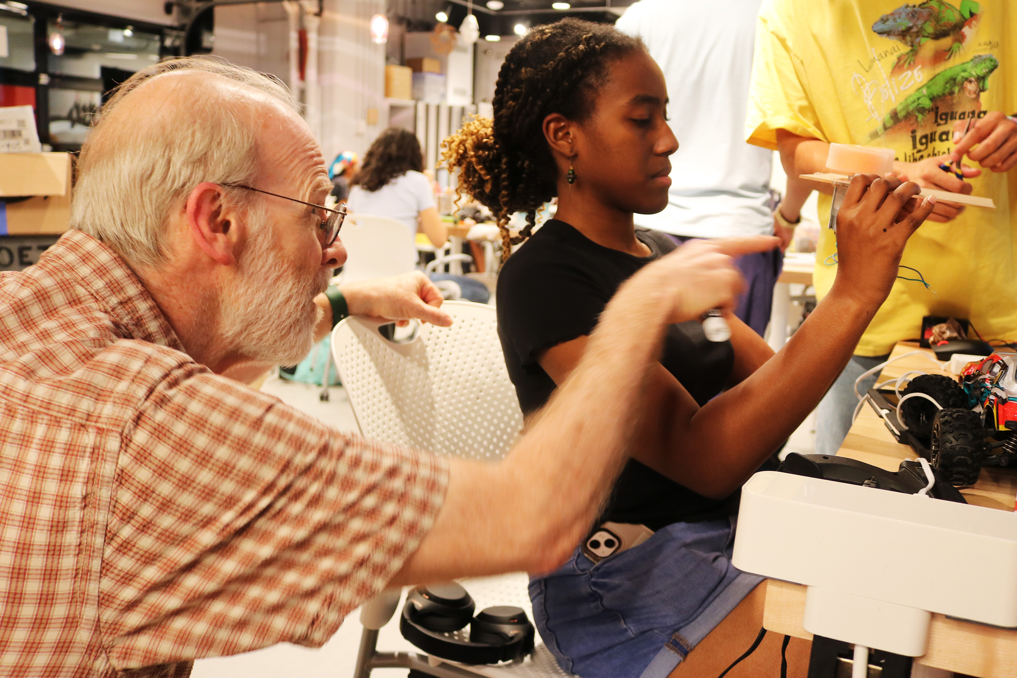 Ed Moriarty points at a piece of wood a student is holding. She is seated at a table where toy car parts and a glue gun are visible.