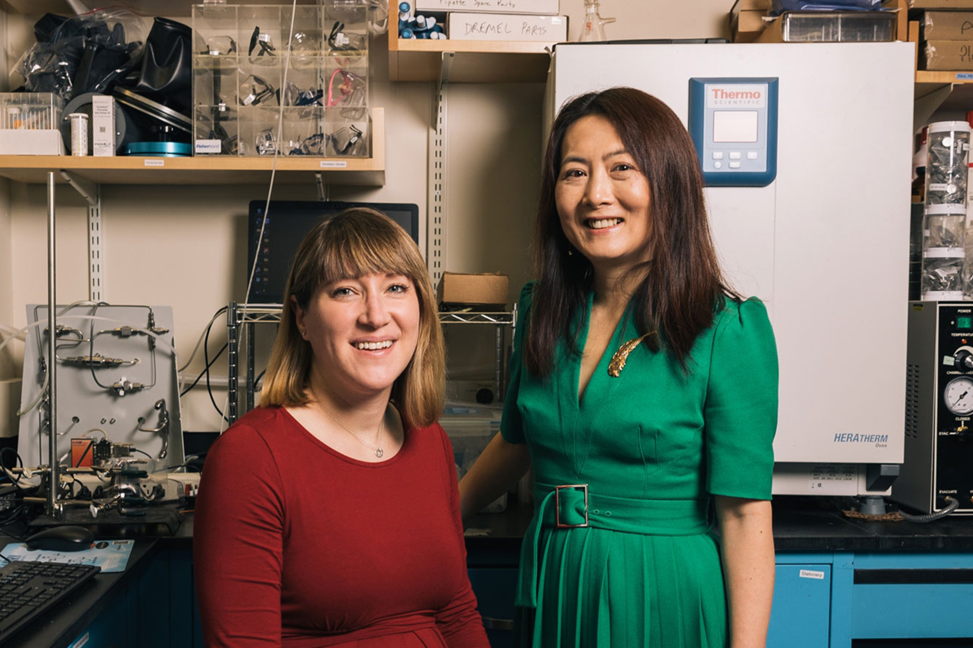 Betar Gallant, left, and Yang Shao-Horn pose in a lab. Gallant is seated and wears a red dress. Shao-Horn is standing and wearing a green dress. Shelves can be seen in the background, and a small refrigerator.