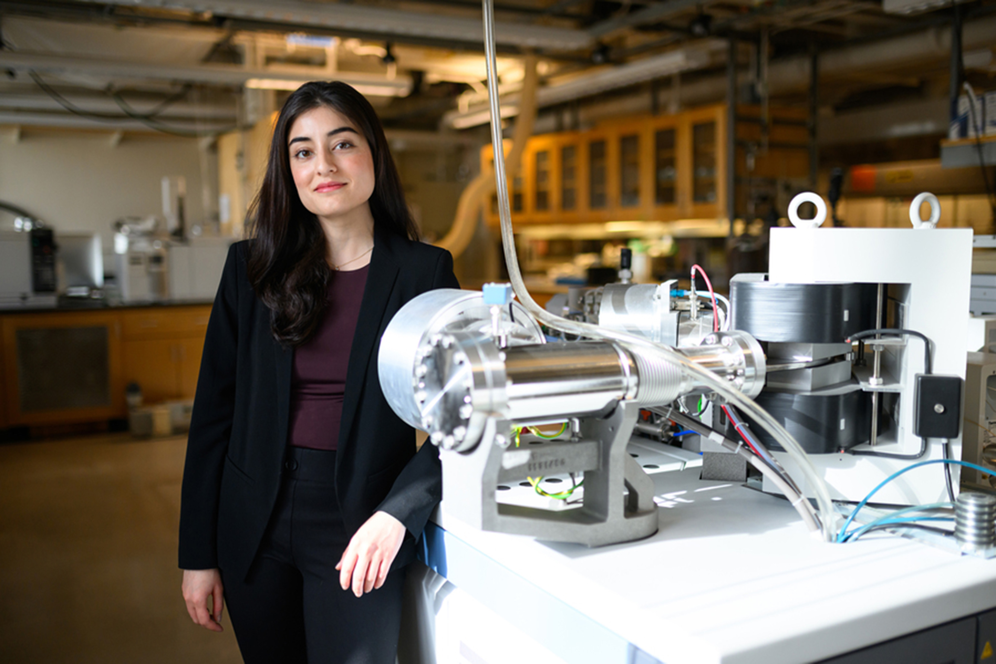 Fatima Husain stands in a lab with arm resting on table. The table has a complex machine with a cylindrical base and tubes and wires connected to the table and ceiling.