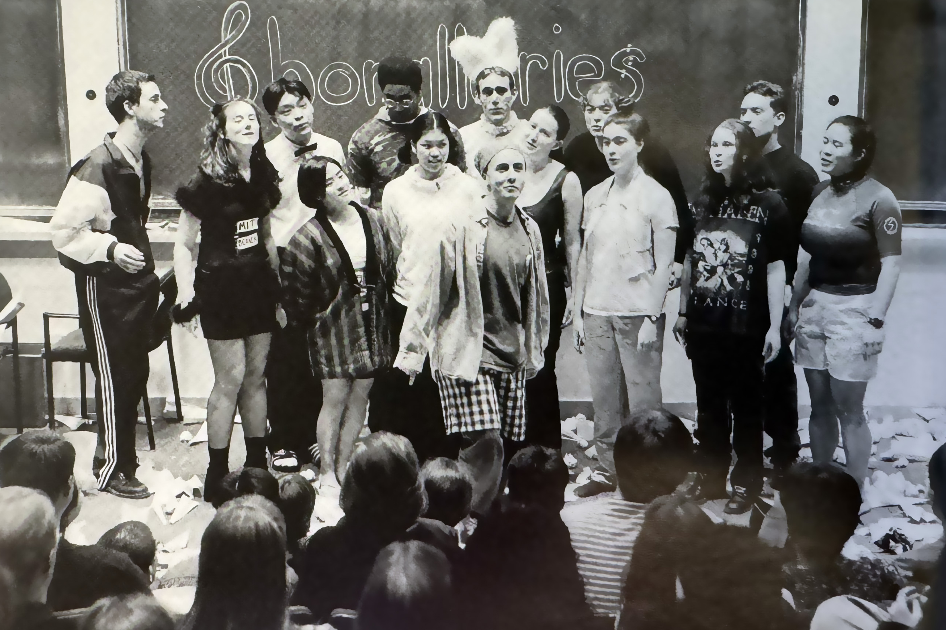 An old black and white photo shows a group of singers in front of a blackboard on which the word Chorallaries is written. The singers are wearing an assortment of garments, including a feather headress, a bandana, and a bowtie. The floor is littered with trash. The heads of some audience members appear in the foreground.