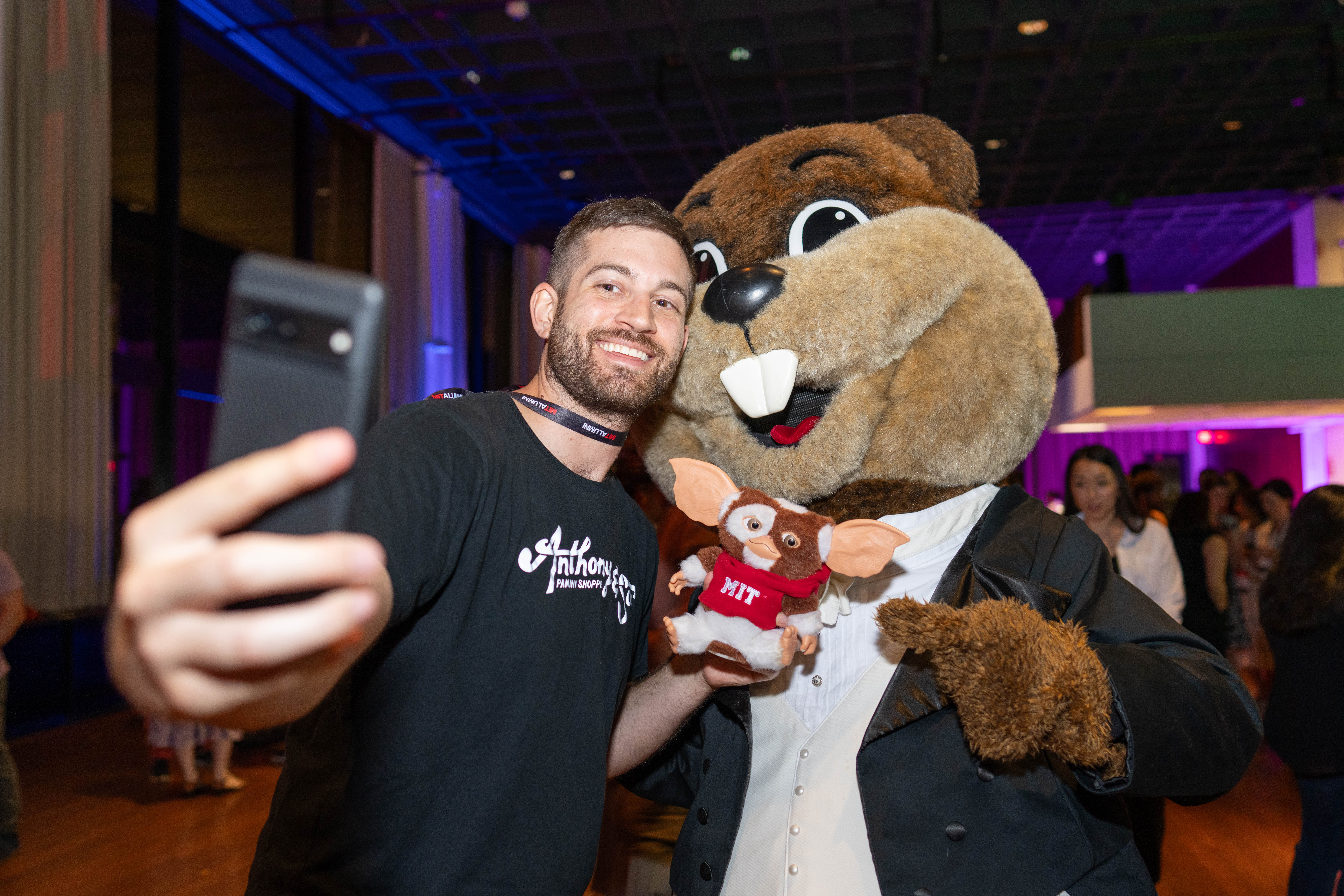 A man in a black T-shirt takes a selfie with Tim the Beaver. Some people are visible in the dark background.