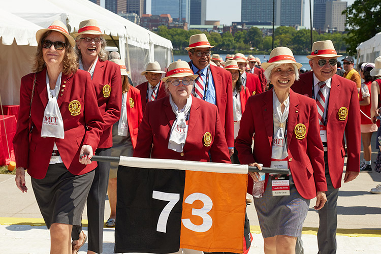 Three people in red MIT jackets and straw hats lead a procession while holding a black and orange 73 banner. The river and some Boston buildings can be seen in the background.