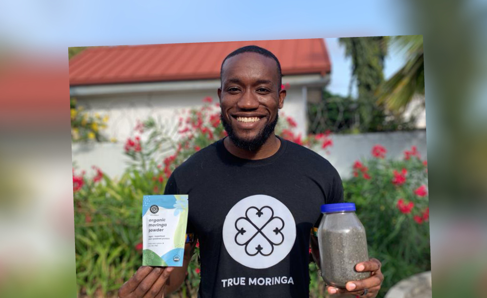 Kwami Williams standing outside with a white building behind him with a red roof, holding True Moringa products and wearing a True Meringa tshirt 