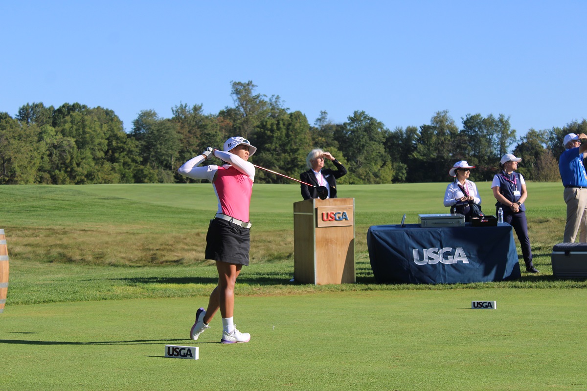 A photo of MIT alum Kimberly Dinh swinging a golf club outside on a golf course with a person behind her at a podium