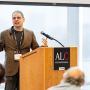 Professor Aleksander Mądry gestures with open hands behind a podium labeled ALC. He wears a brown jacket and an ALC badge.