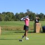 A photo of MIT alum Kimberly Dinh swinging a golf club outside on a golf course with a person behind her at a podium