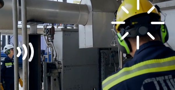 Two workers are shown wearing helmets and ear protection in an industrial setting. White marks overlaying the image indicate broadcasting from one and receiving by the other.