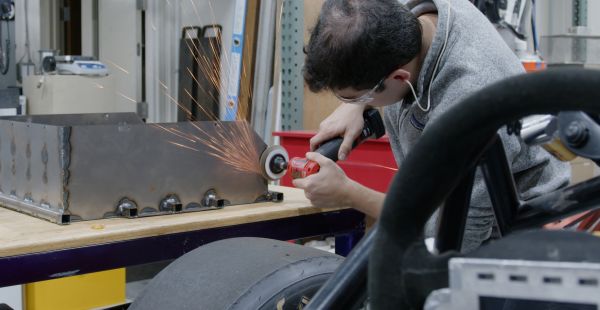 A man leans over a workbench holding a grinder against a metal surface. Sparks fly. A tire and a steering wheel are visible in the foreground.